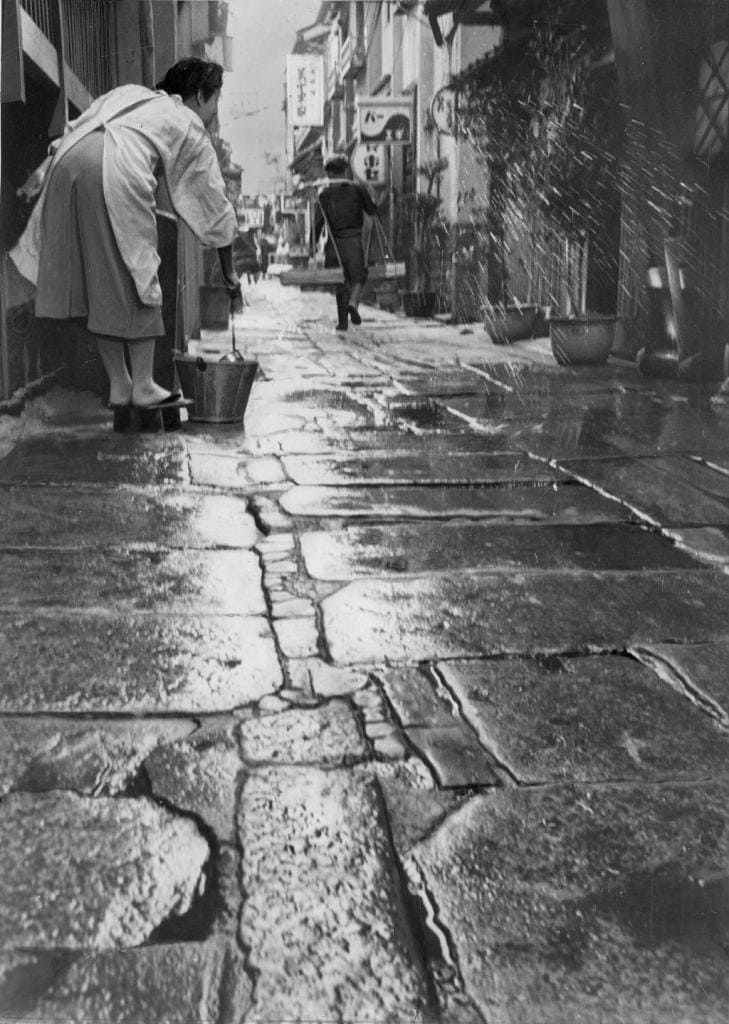 1955（昭和30）年の大阪・千日前の朝の法善寺横丁。太陽が昇りきる前に打ち水をするのが、各商店の日課。奥には天秤を担いだ物売りの姿が見える