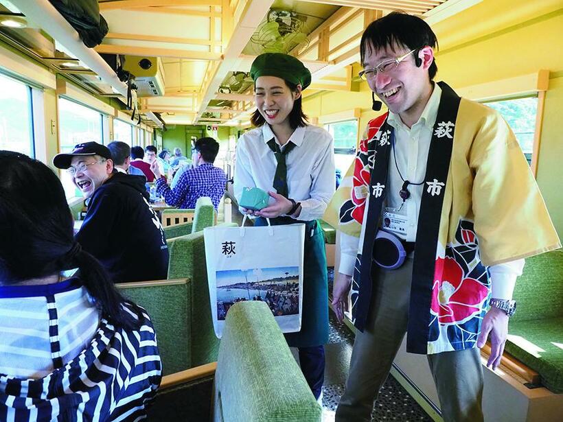 和風インテリアの１号車でクイズの賞品、萩焼の湯のみをプレゼントする萩市観光課の山谷義貴さん（右）とアテンダントの峰添晴子さん。この日の「〇〇のはなし」は、萩がテーマだったが、運行日によって、「川棚温泉のはなし」「鉄道のはなし」「焼き鳥のはなし」などが実施される（撮影／櫻井寛）
<br />