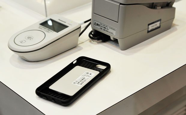 黒いケースの中央に置かれているグレーのカード型をした端末が「おサイフケータイ ジャケット０１」。この上にｉＰｈｏｎｅを装着して持ち歩くことも可能だ。ケースも同時に発売される