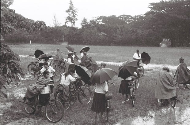 突然の雨。慌てて雨かっぱをかぶったり、傘を差したりして、自転車にまたがり、家路を急ぐ女子学生たち。沖縄本島北部にあった県立第三女学校（通称・三高女、現・名護高校）の生徒と思われる　（C）朝日新聞社