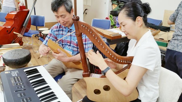 早期認知症治療の一環である音楽治療。山本氏は最近、写真の古楽器「プサルタリー」の演奏にはまっているという。右は、先生の折山もと子さん
