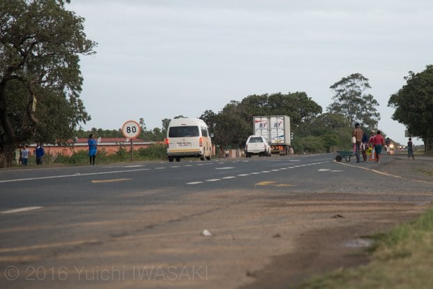 帰宅するためにヒッチハイクをする人が立ち始めた、道路の分岐点。左に見えるワンボックスカーは、乗り合いバス。一般車両でも、空きスペースがあれば人を乗せることは多い（ムクゼ近郊・南アフリカ／Mkuze,South Africa 2016）