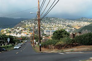 オアフ島の大きな丘のような住宅街。下り坂道の向こうの斜面にはぎっしりと家が立ち並び、どんよりとした雲がおおっている。画面に縦横に入れ込んだ電柱と電線が遠近感を出し、構図のアクセントとなっている