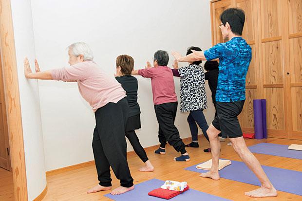川村さんの提唱するひざ裏伸ばし体操の一つをする高齢者ら。手をついて壁を押しながら、ひざ裏の筋肉を伸ばす簡単なストレッチだ