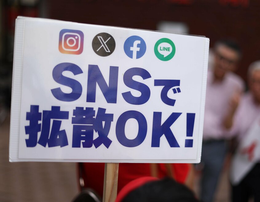 立候補予定者が記念写真を撮るそばで、陣営スタッフが「SNSで拡散OK!」と書かれた看板を掲げていた=2025年5月25日、神戸市中央区