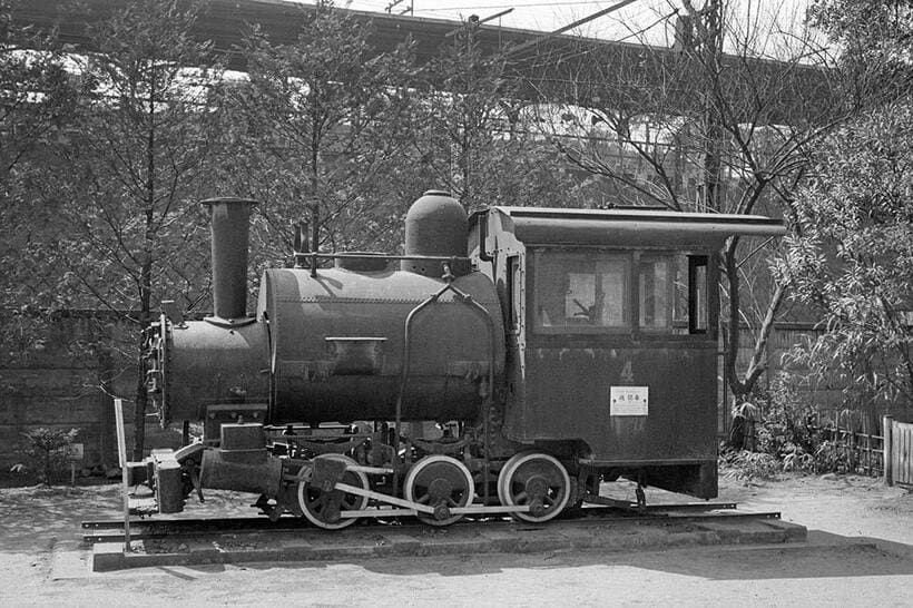 製紙博物館の前庭に展示されていた王子製紙軽便鉄道で使われた4号機関車。ボイラの上に水タンクを載せたサドルタンク仕様の特異な外観。背景に都電の架線と国鉄（現JR）王子駅のホームが見える（撮影／諸河久：1963年4月3日）