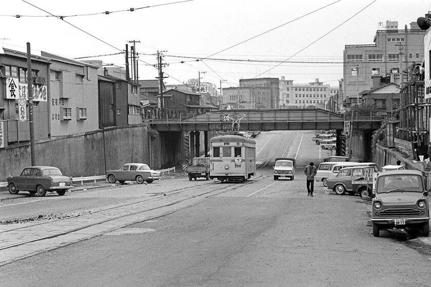 国鉄高島貨物線の跨道橋をくぐり抜けて終点中央市場に向う7系の市電。中央市場通りの左側にはヒルマンミンクス、日産ブルーバード310、右側にはマツダB360などが駐車していた。背景の神奈川会館は老朽化のため1983年に惜しくも解体された。神奈川会館前～中央市場（撮影／諸河久：1965年11月30日）
