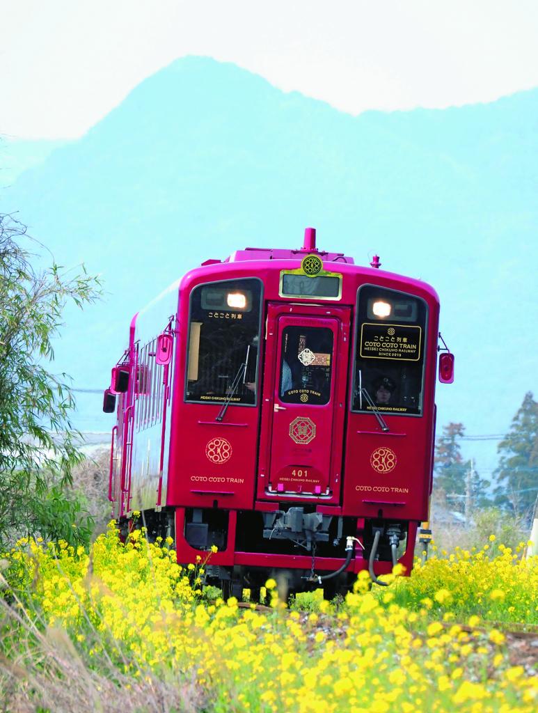 筑豊の田園地帯をコトコト走る平成筑豊鉄道の新鋭「ことこと列車」。菜の花は満開にして、背後の筑豊の山並みは春霞という、いかにも春のうららかな風景の中、レッドメタリックの車体がまぶしい。ローカル線だからこそより華やかにという水戸岡デザイナーの色彩感覚が光る。数多い赤い列車の中でも最もあでやかな赤ではないだろうか。田川線豊津駅─今川河童駅間で　■オリンパスOM－D E－M1 MarkII・40～150ミリ F2.8＋TC1.4・絞りf8・ISO800・AE・マイナス0.3補正・JPEGスーパーファイン